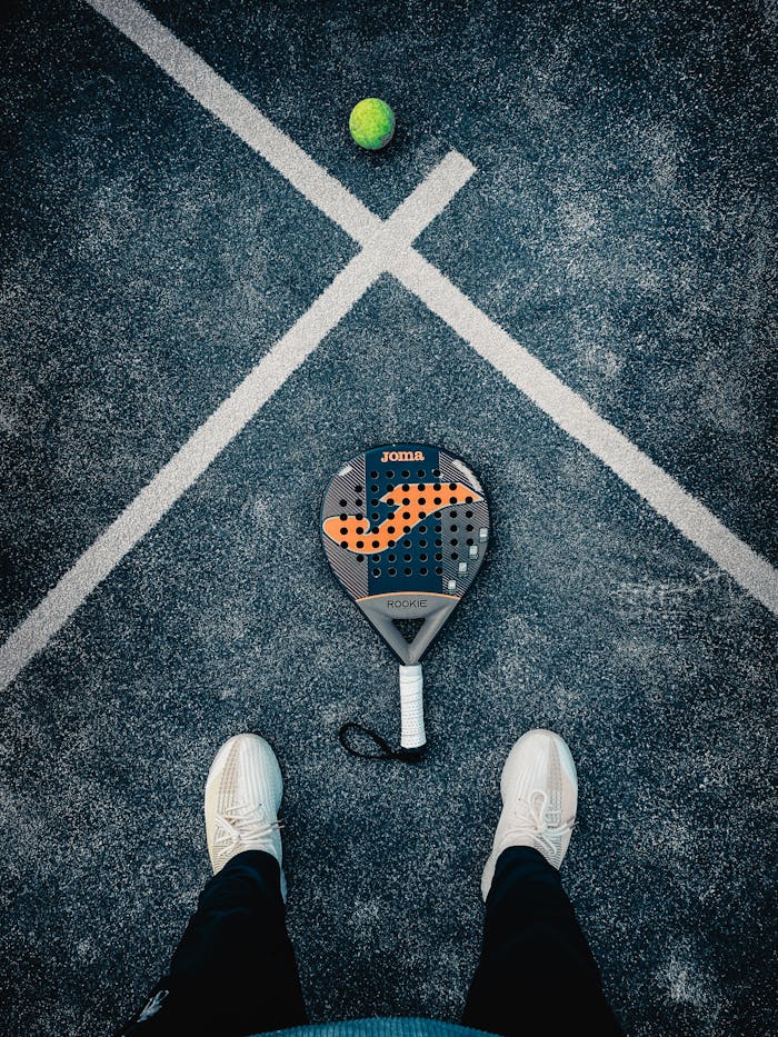 Flat lay of a padel racket, ball, and shoes on textured court surface.
